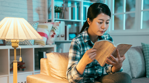 Woman sitting in living room reading a book by a lamp